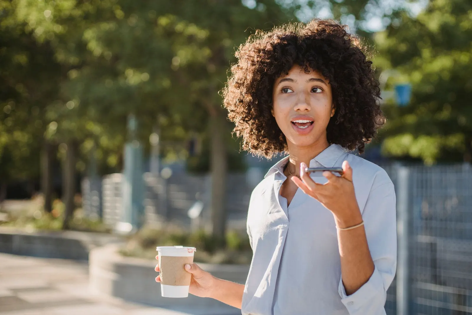 A woman speaking into her phone outdoors with a coffee in hand