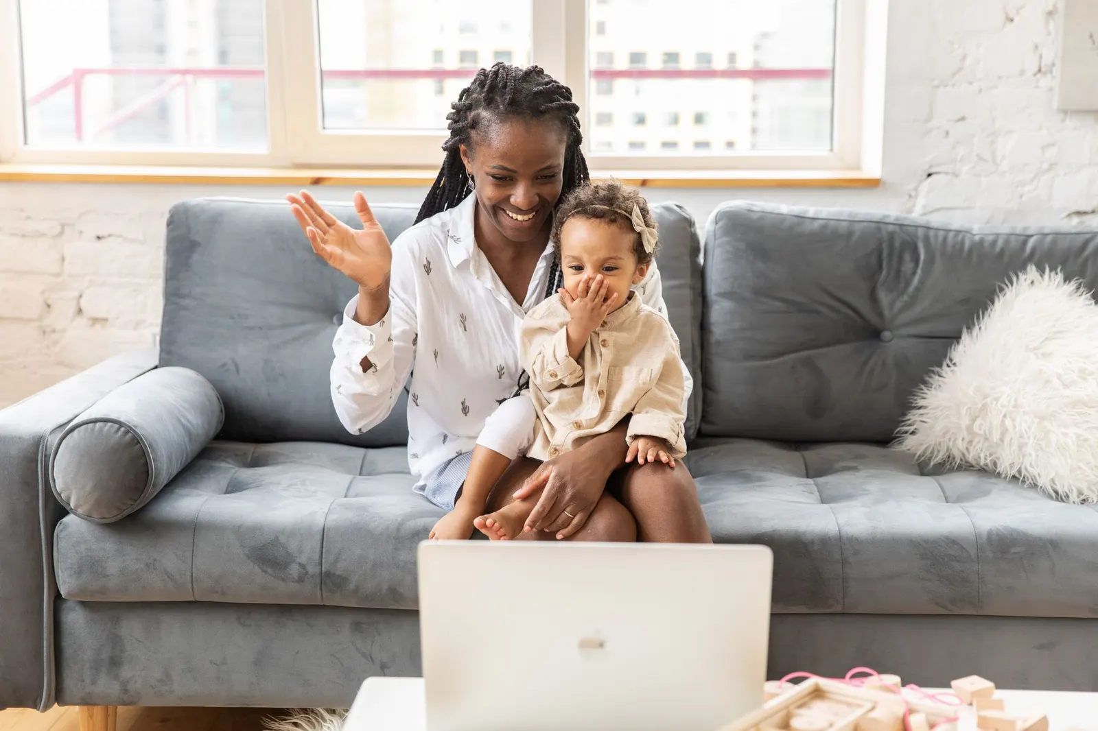 A mother and her little daughter waving to family on a video call from the sofa