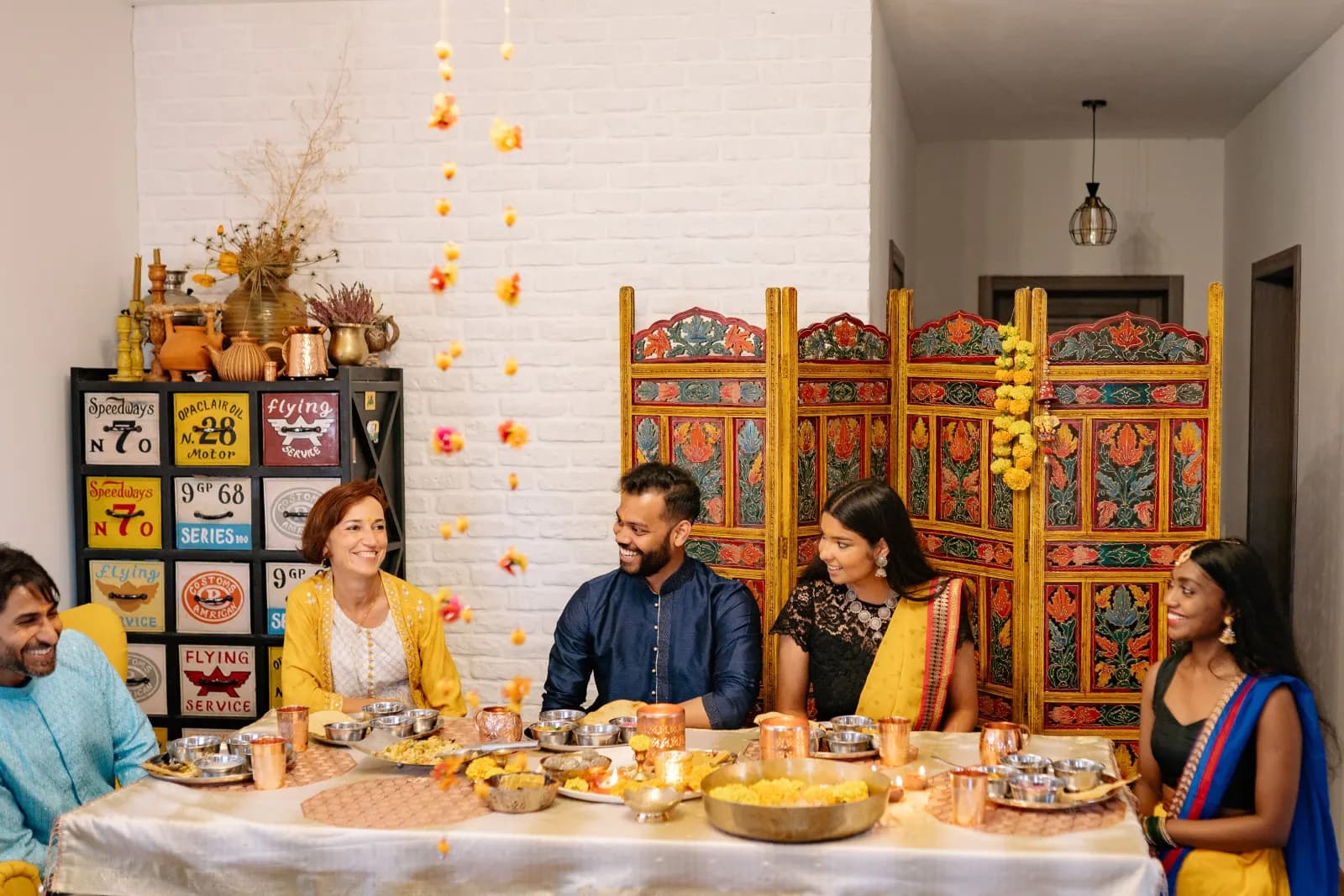 Family members laughing together around a table set for a Diwali meal, with lamps and marigolds