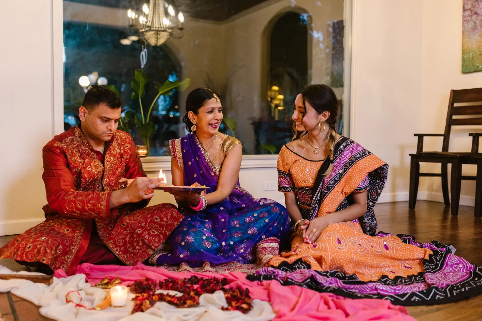 Three adults sitting on the floor in festive clothes, lighting a diya from a puja plate
