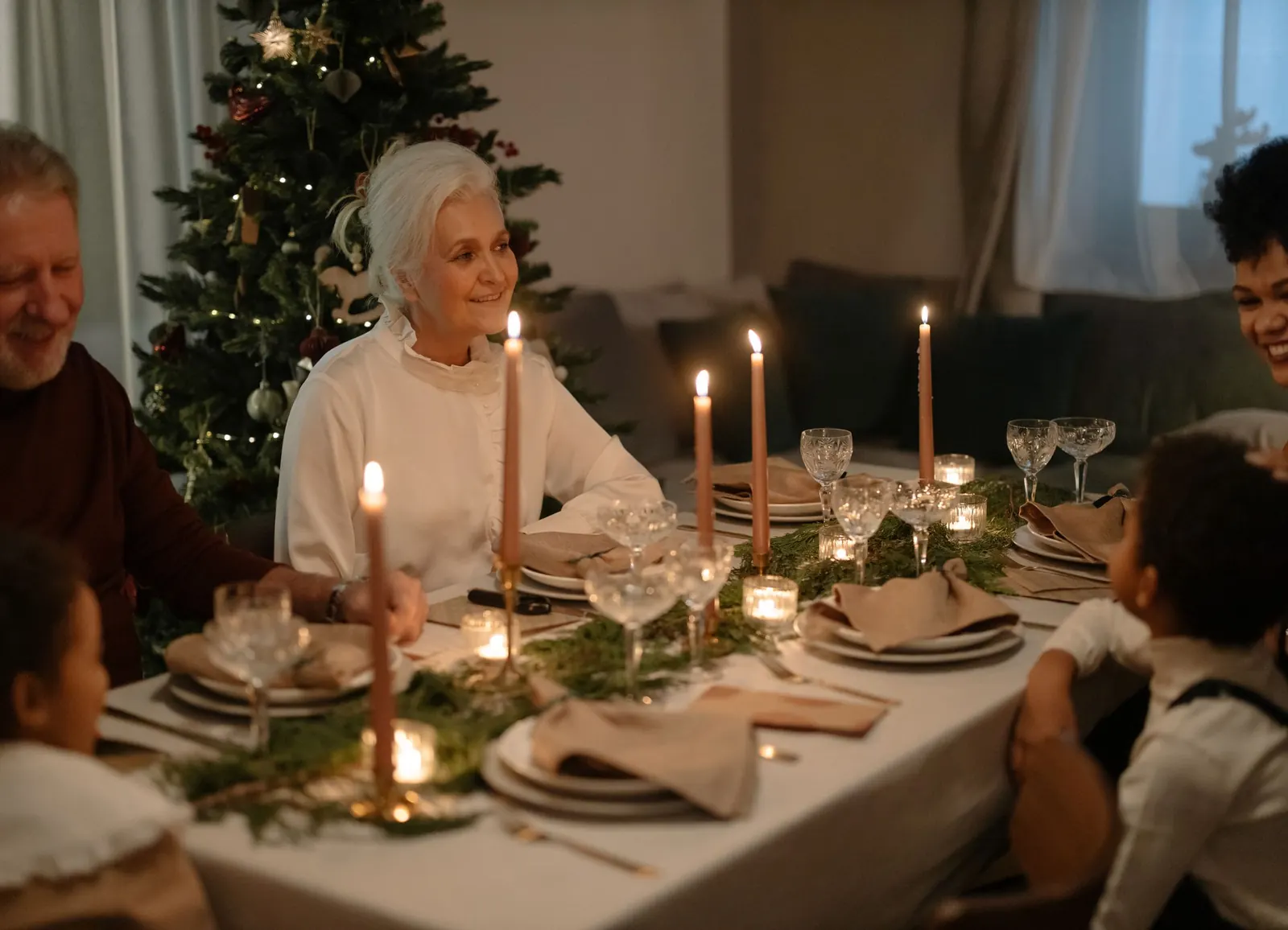 An older woman smiling at a candlelit Christmas dinner table with her family, an evergreen tree behind her